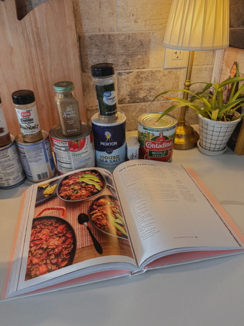 cookbook displaying chili recipe and ingredients sitting on kitchen counter. There's also a plant, wood serving board, and small lamp.